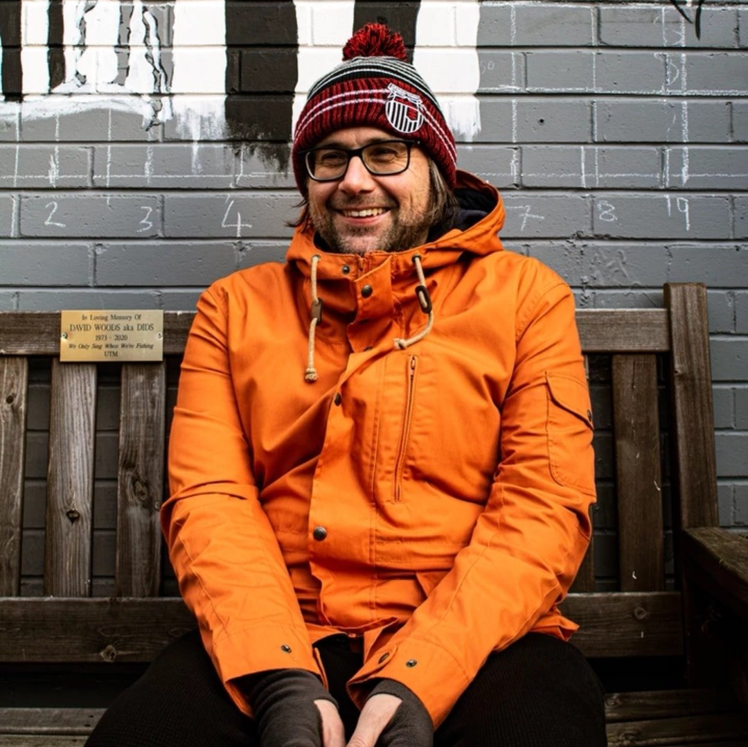 A man in an orange jacket and hat is sitting on a bench