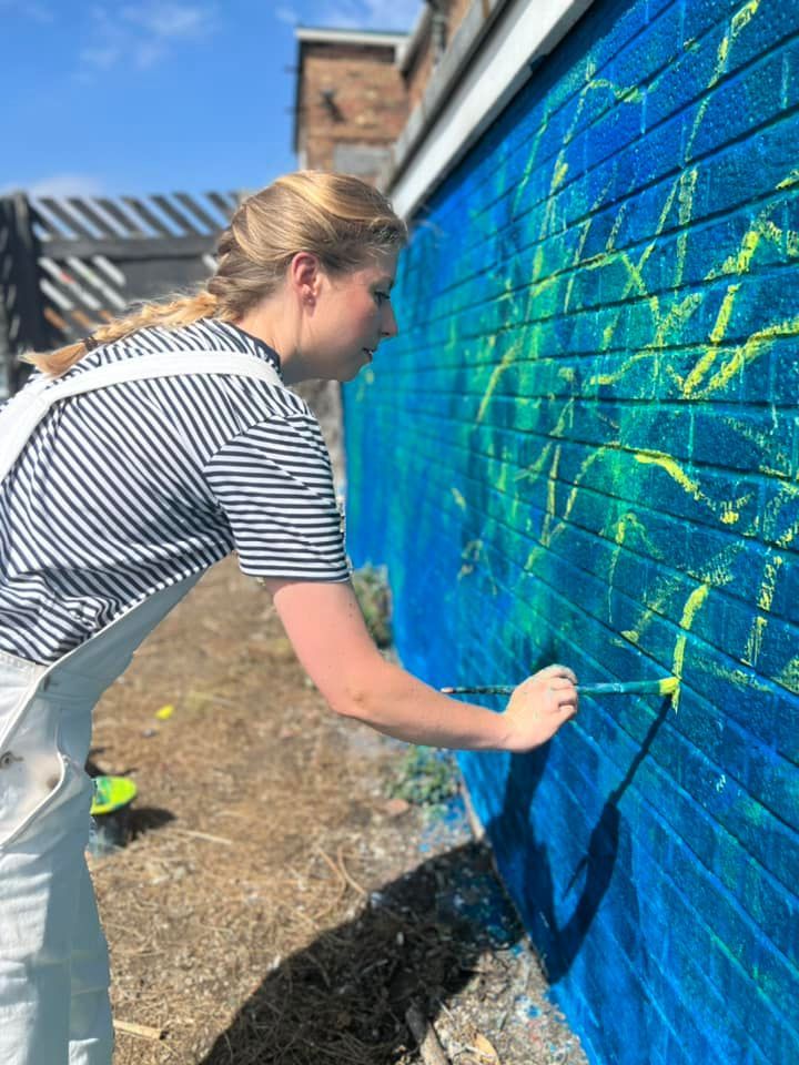 A woman is painting a mural on a blue brick wall.