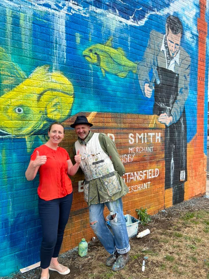 A man and a woman are standing in front of a mural on a brick wall.