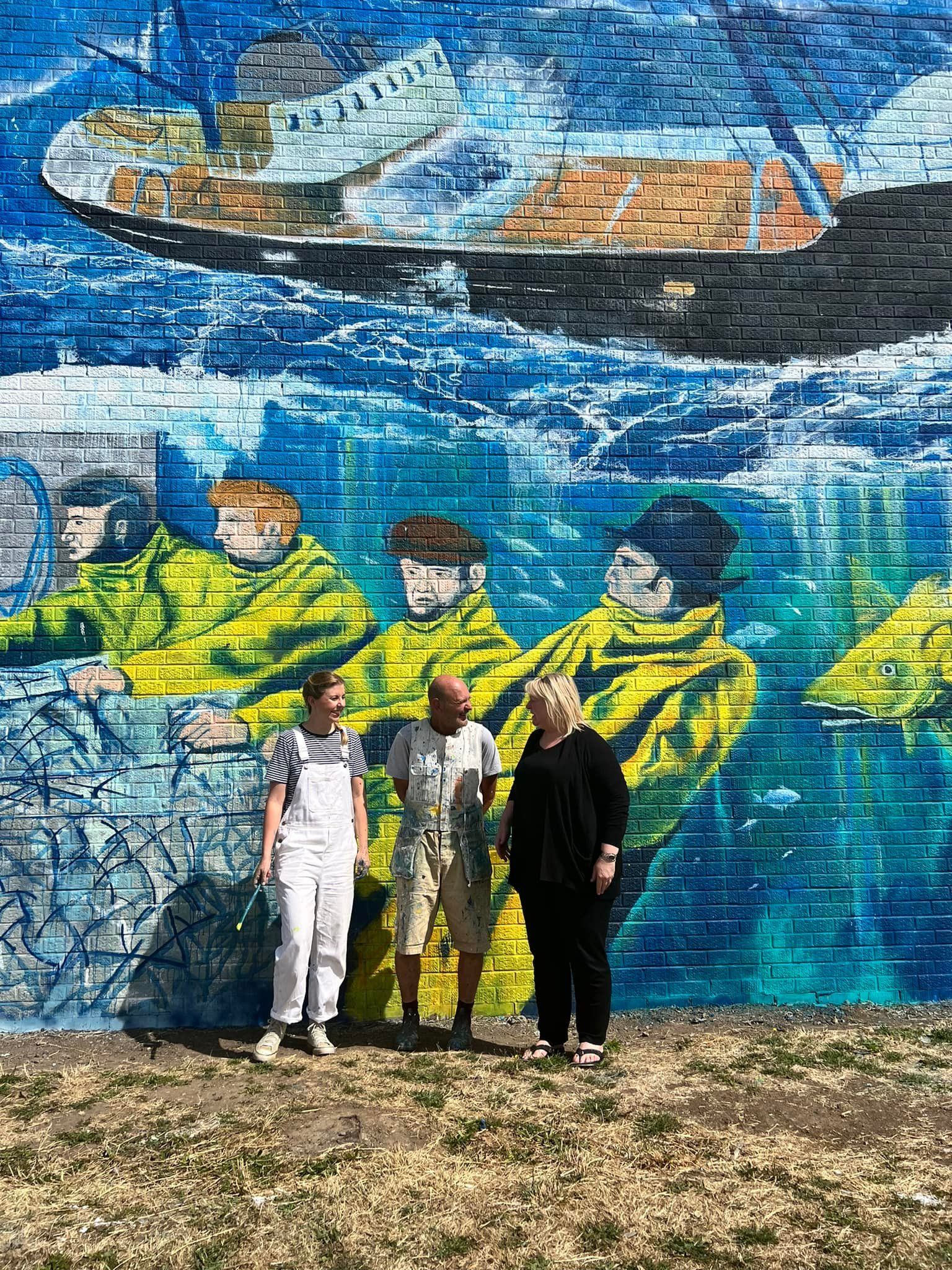 Three people are standing in front of a large mural on a brick wall.