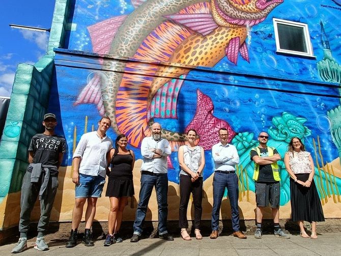 A group of people are standing in front of a mural of a fish on a building.