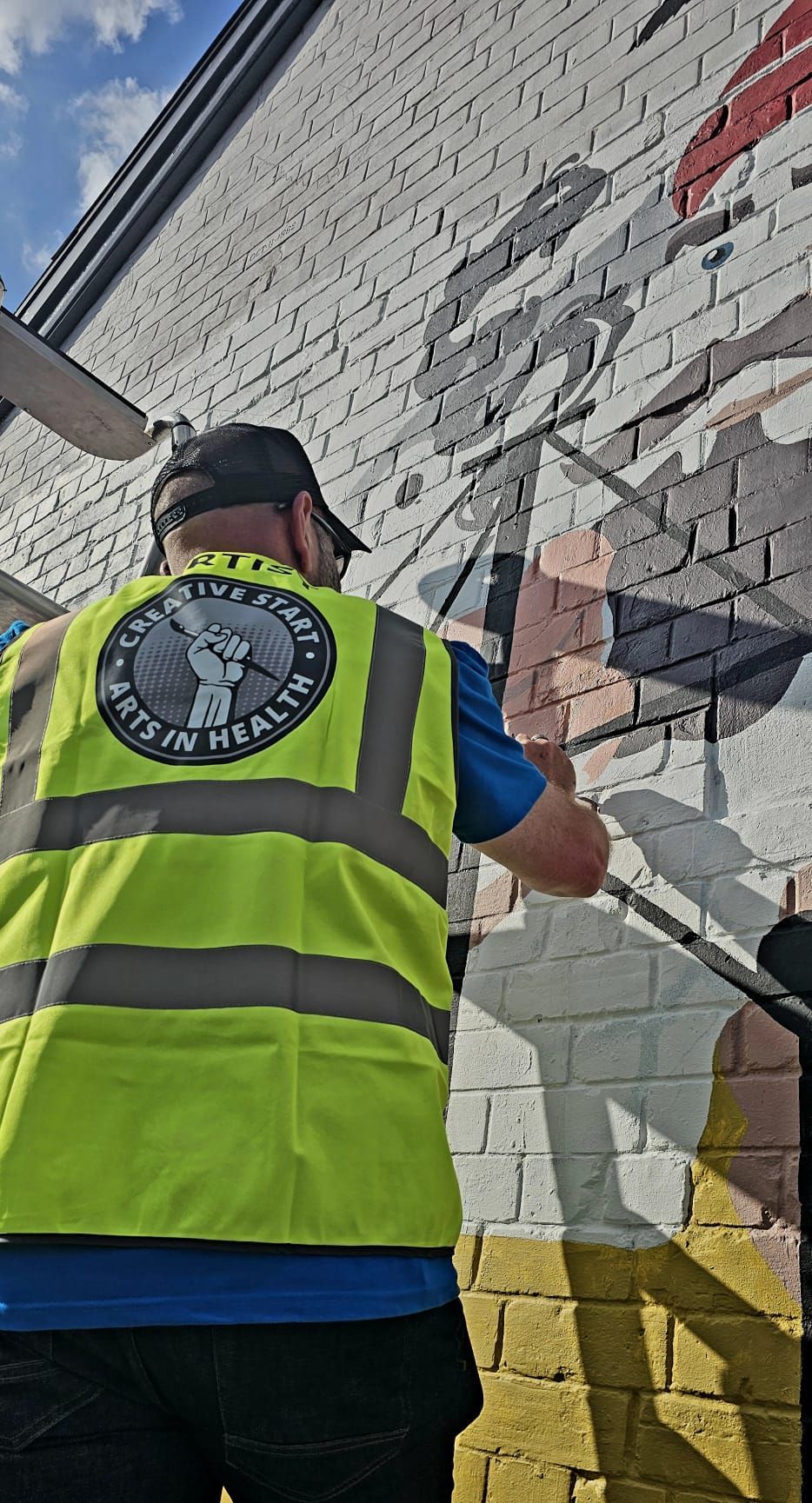 A man in a yellow vest is painting a mural on a brick wall.