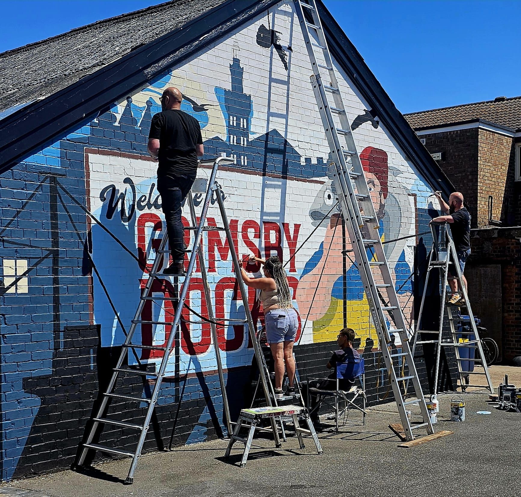 A group of people are painting a mural on the side of a building that says welcome grimsby