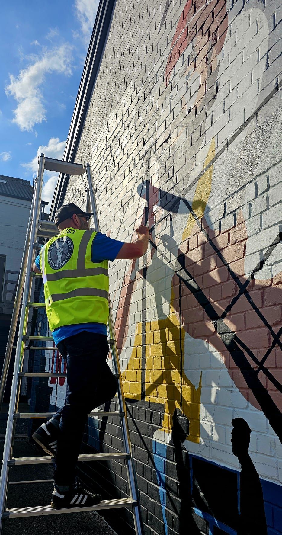 A man is standing on a ladder painting a mural on a brick wall.