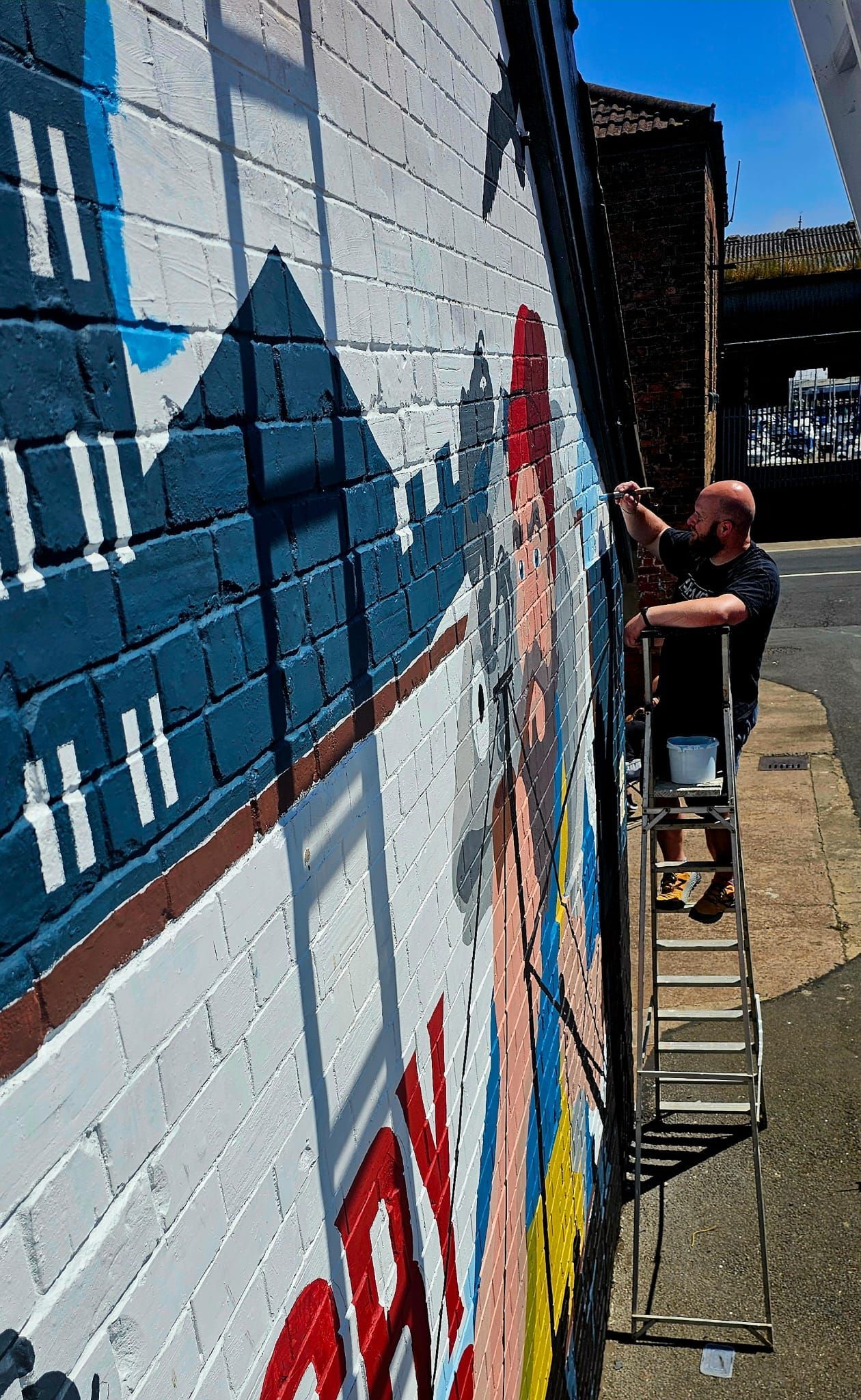 A man is standing on a ladder painting a mural on a brick wall.