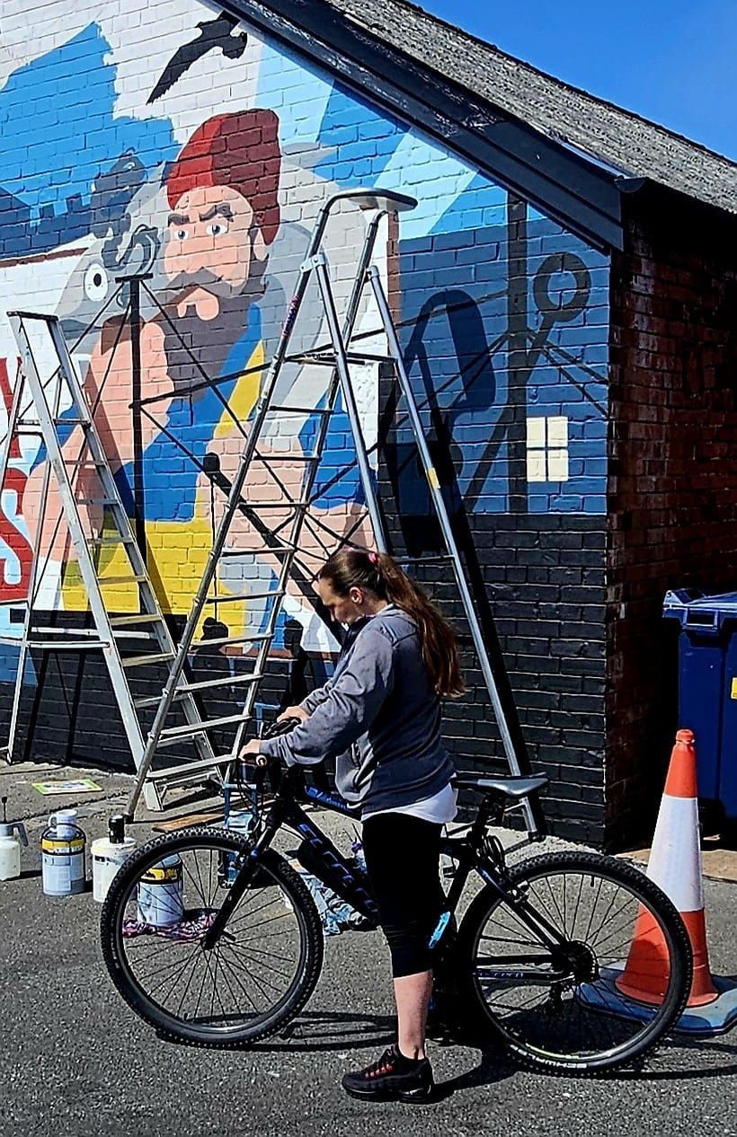 A woman is standing next to a bicycle in front of a mural on a wall.