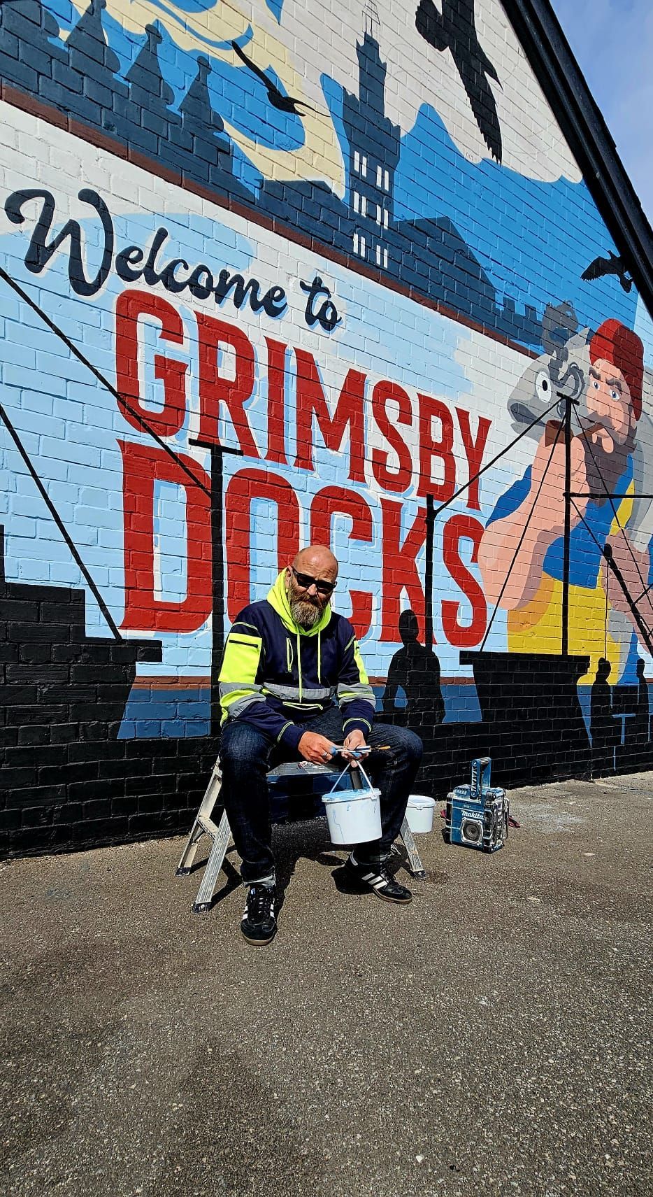 A man is sitting in front of a mural that says welcome to grimsby docks.