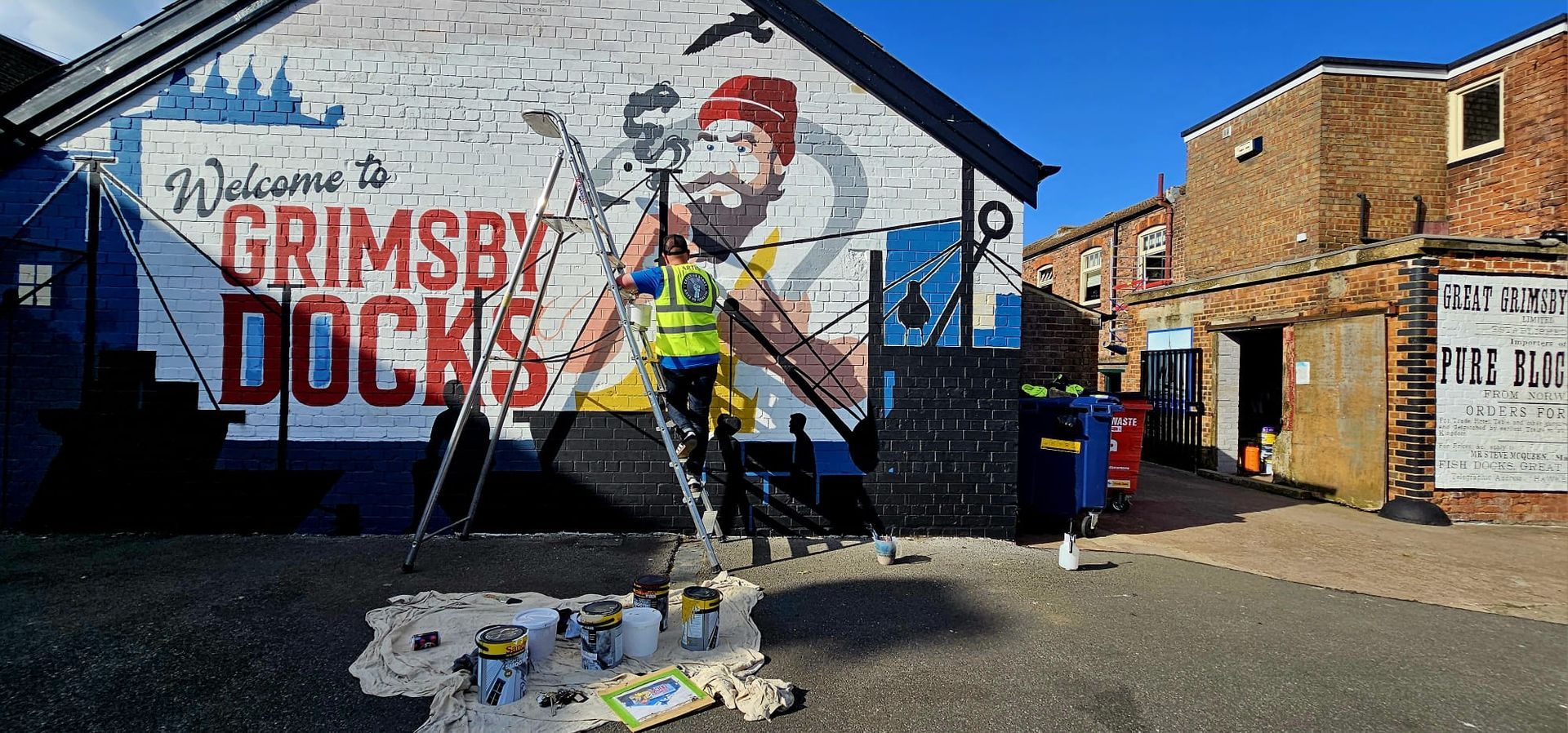 A man is painting a mural on the side of a building that says grimsby docks.