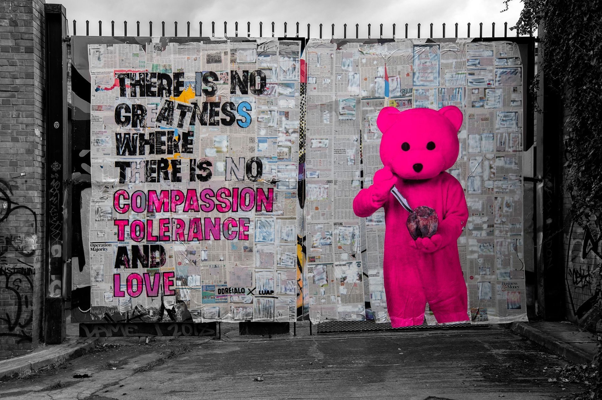 A pink teddy bear is standing in front of a brick wall with graffiti on it.