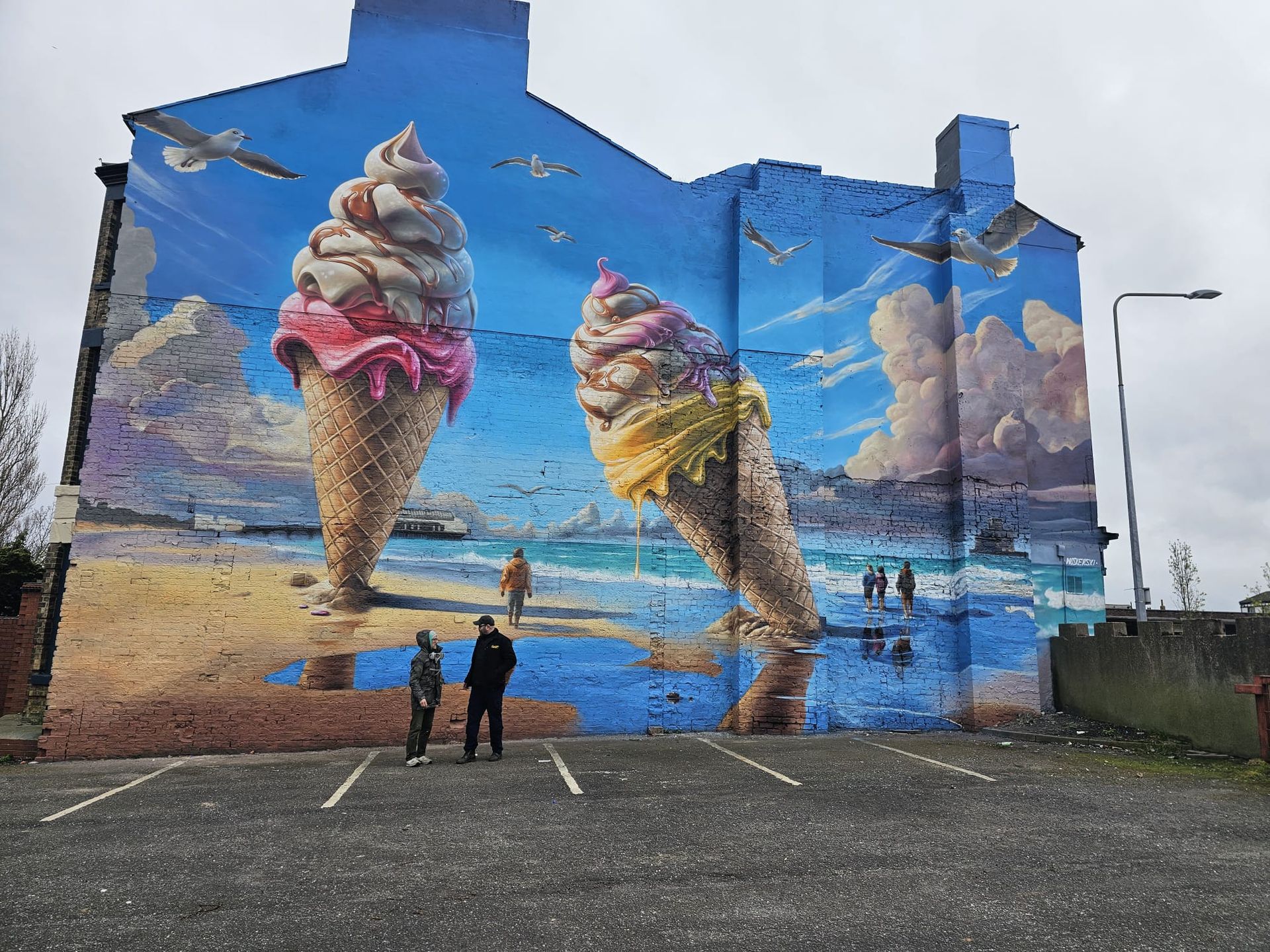 Two people are standing in front of a large mural of ice cream cones on the side of a building.