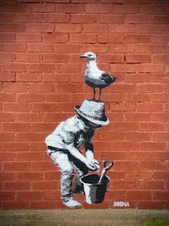 A boy is playing with a bucket and a seagull is sitting on his hat.
