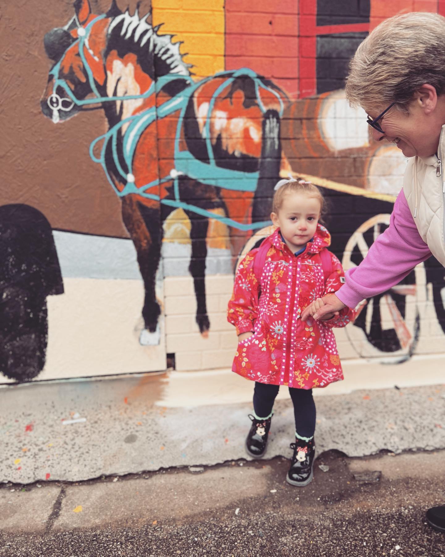 A little girl in a pink jacket is holding a woman 's hand in front of a horse drawn carriage mural