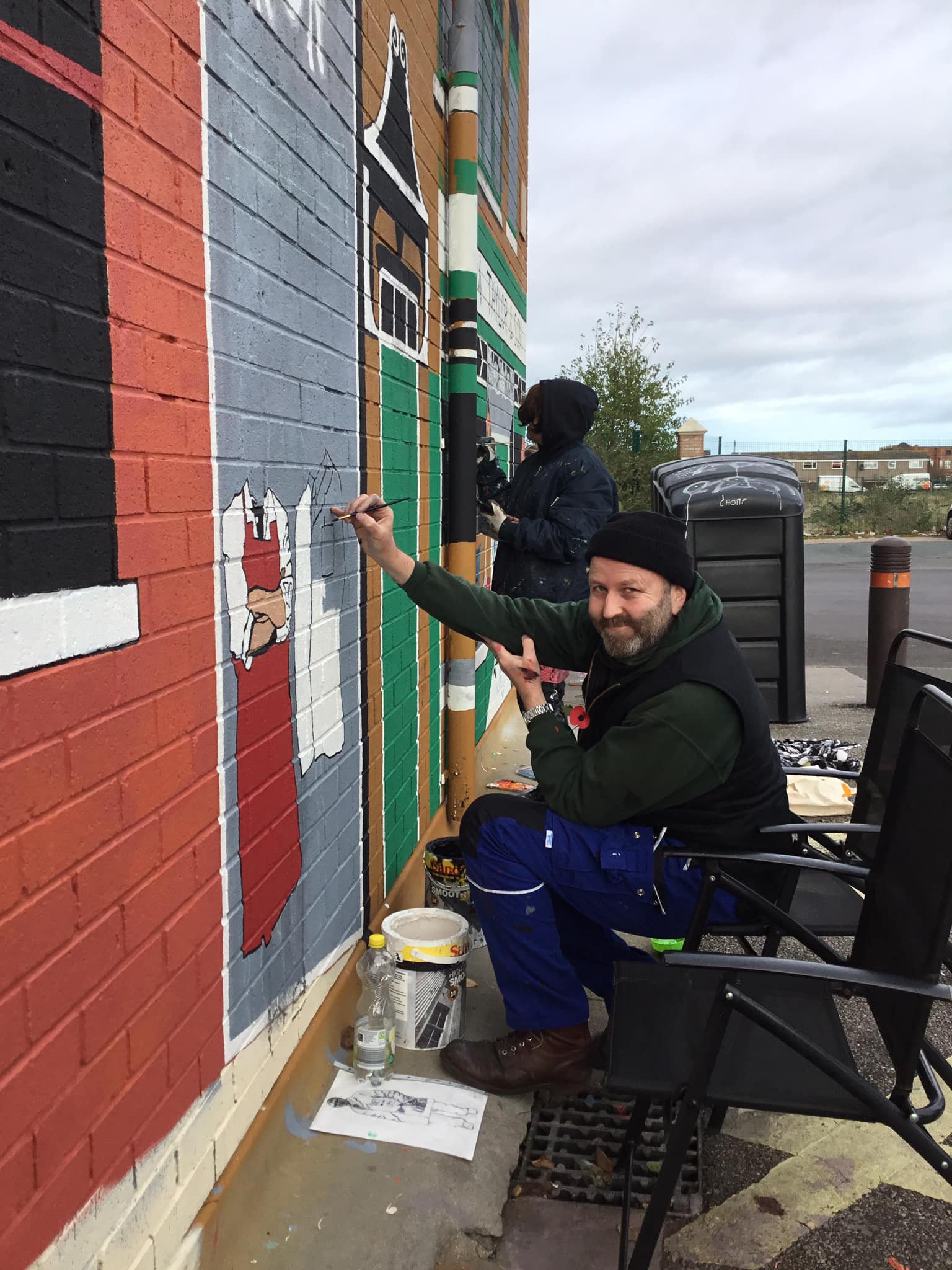 A man is sitting in a chair painting a mural on a brick wall.