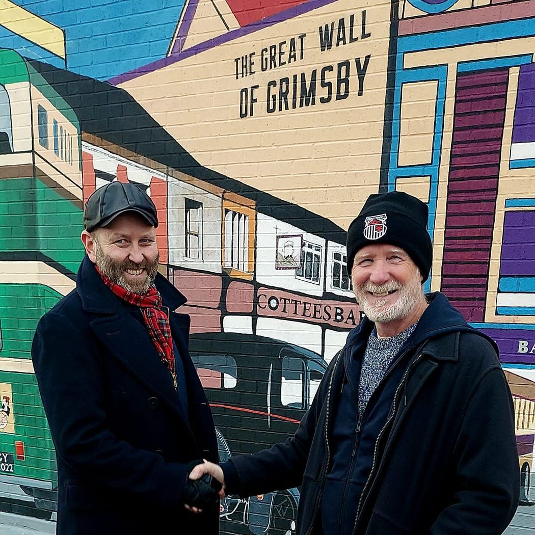 Two men shaking hands in front of a mural that says the great wall of grimsby