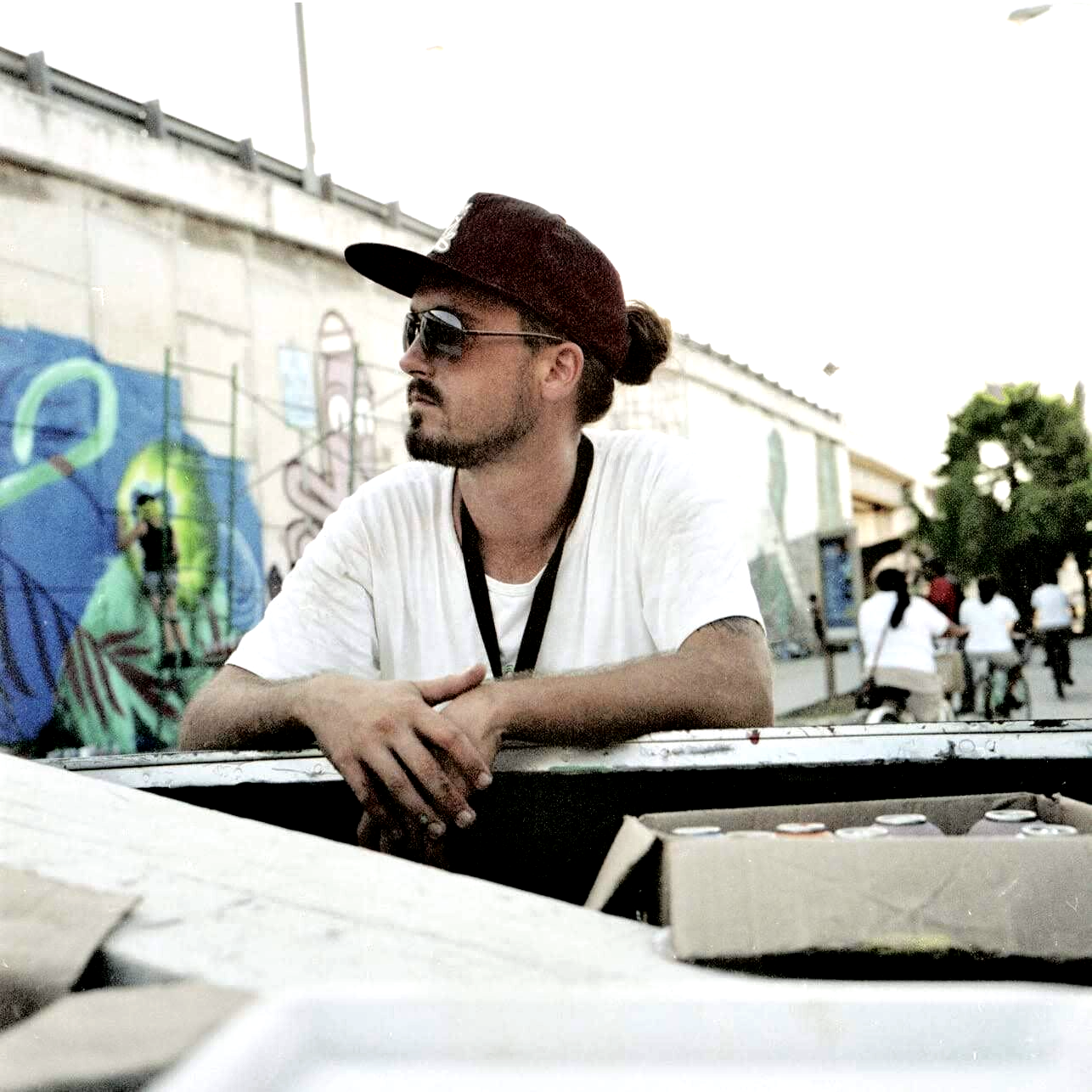 A man wearing sunglasses and a hat leans against a fence