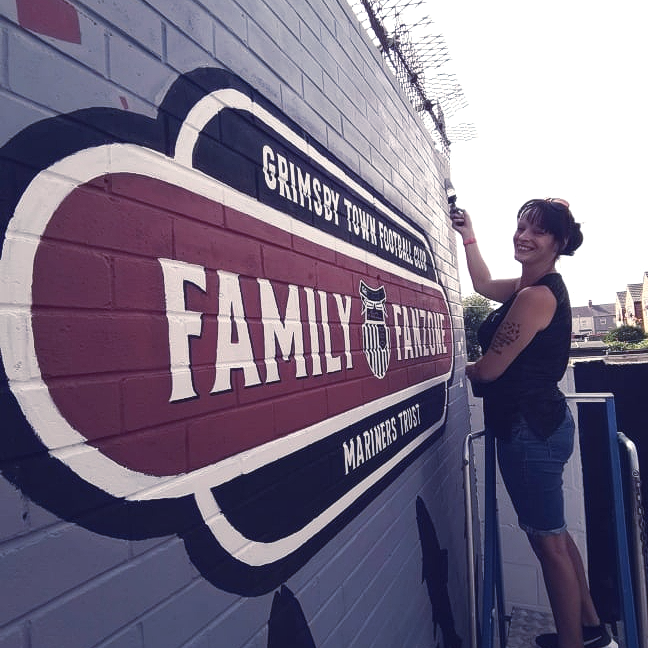A woman paints a sign on a brick wall that says grimsby town football club