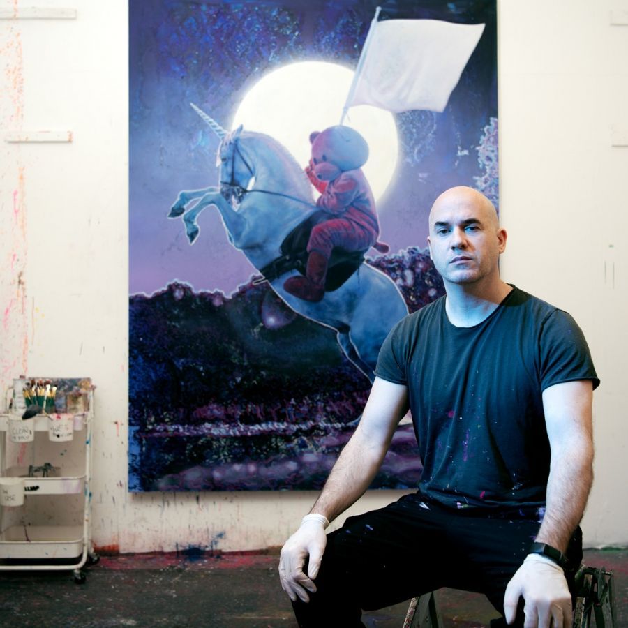 A man in a wheelchair is sitting in front of a painting of a teddy bear riding a unicorn