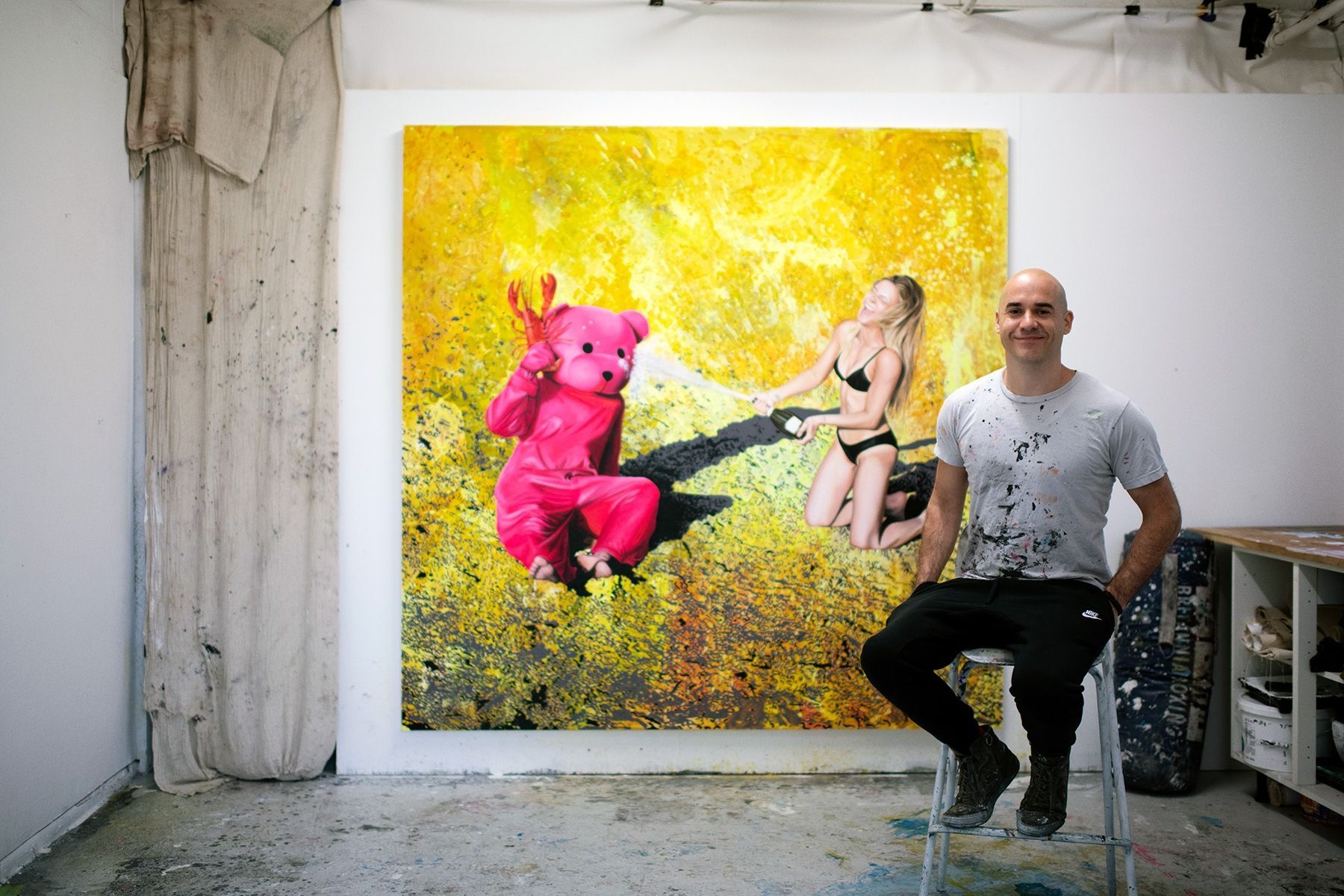 A man is sitting in front of a painting of a woman and a pink teddy bear.