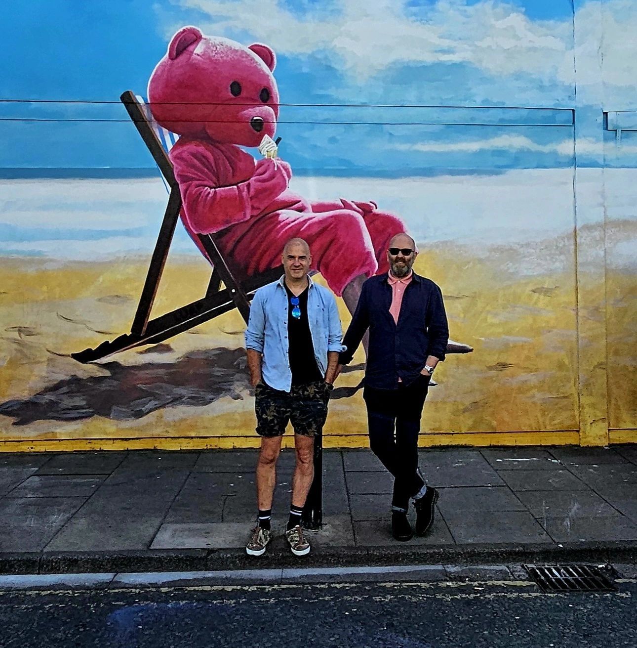 Two men standing in front of a mural of a teddy bear on the beach