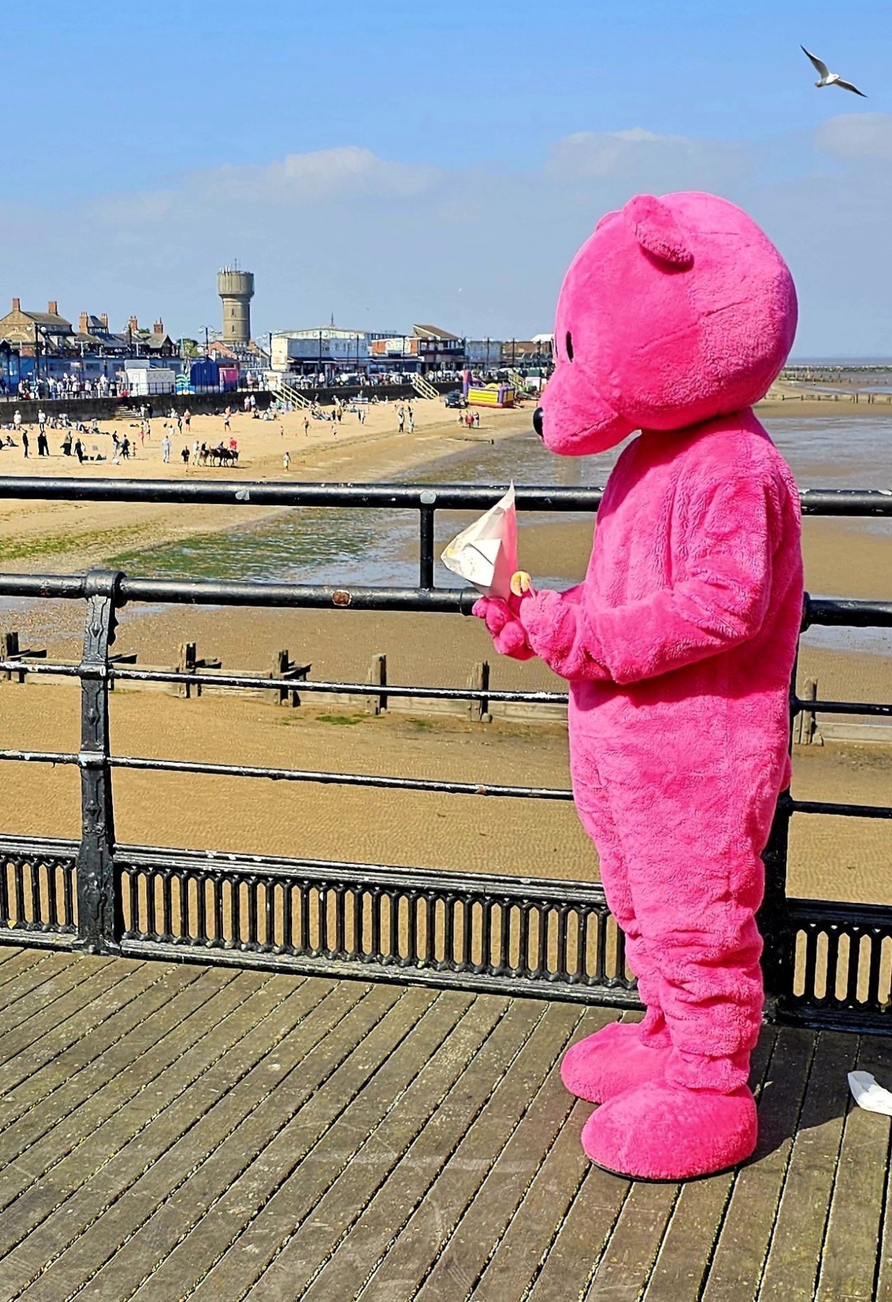A pink teddy bear is standing on a pier holding a piece of paper