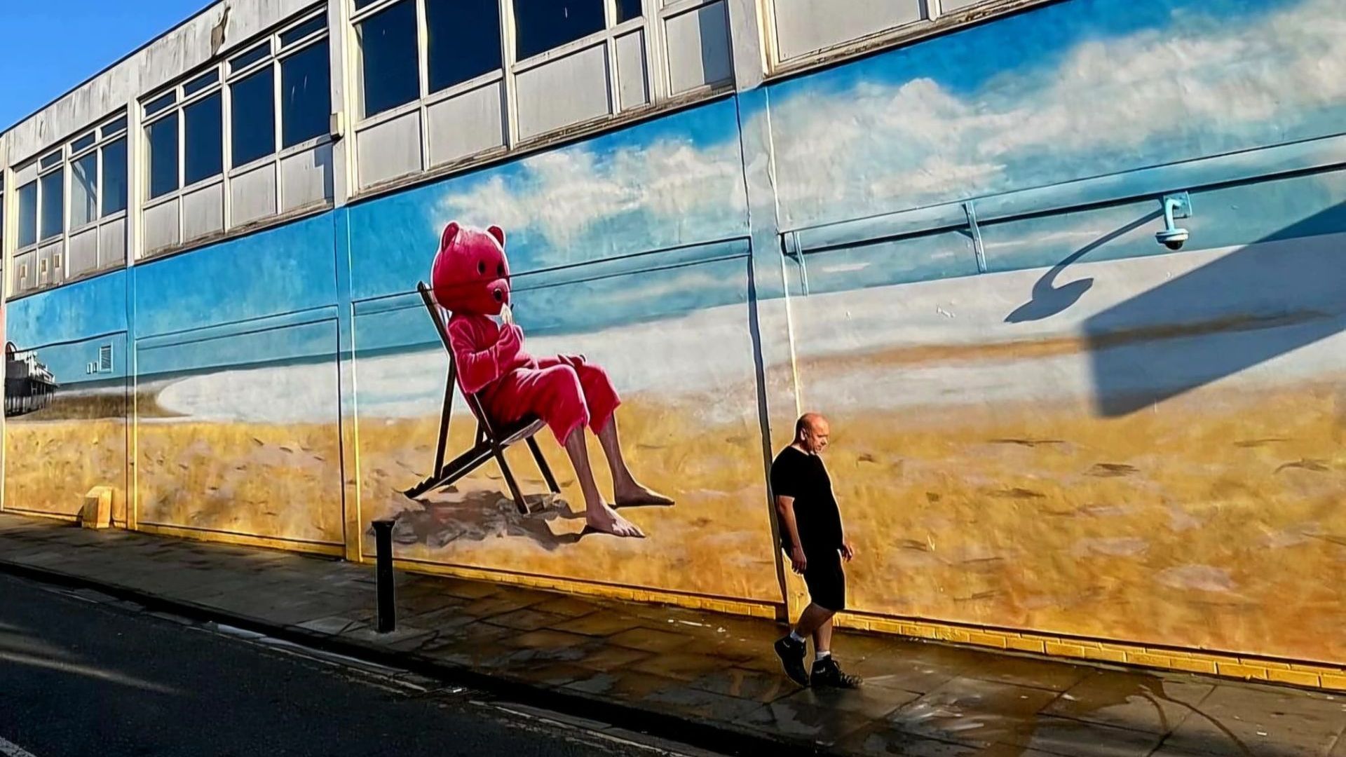 A man is walking down a sidewalk in front of a large mural of a pink teddy bear on a beach.