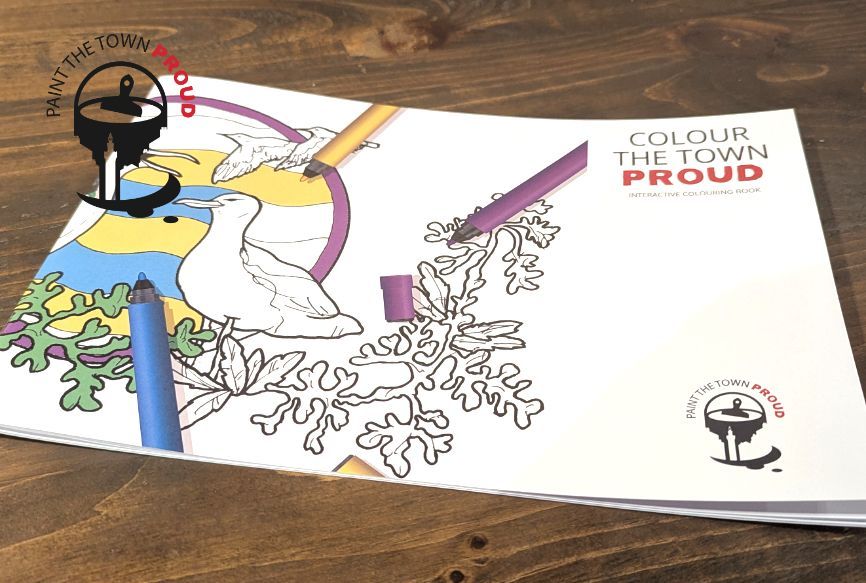 A coloring book titled colour the town proud is sitting on a wooden table.