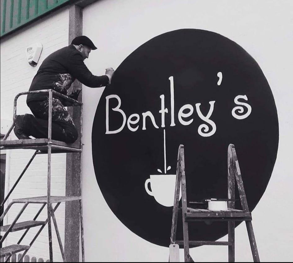 A man paints a bentley 's sign on the side of a building