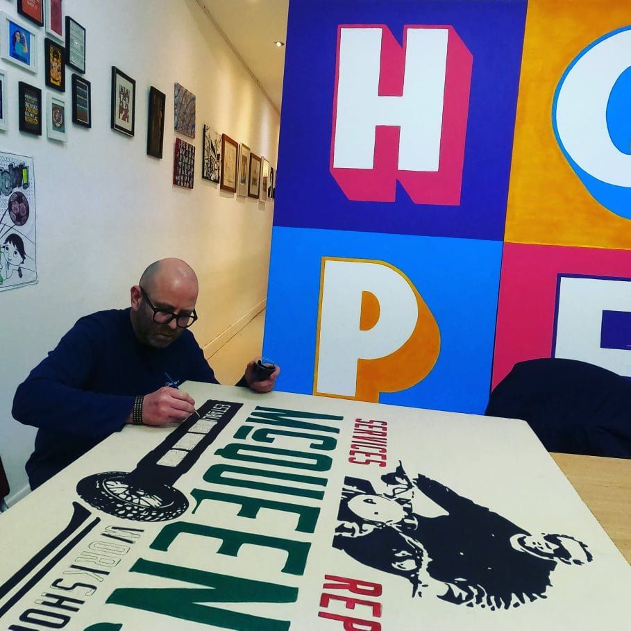 A man sits at a table with a sign that says mcqueens workshop