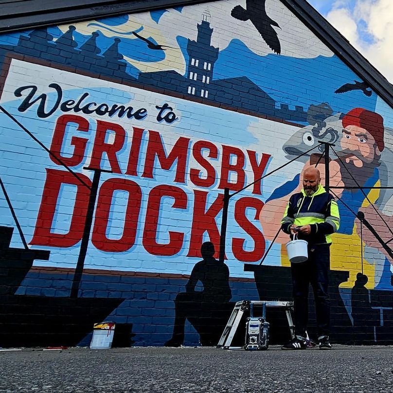 A man is standing in front of a mural that says welcome to grimsby docks