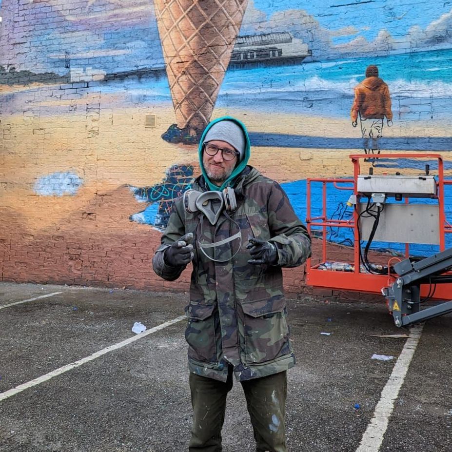 A man in a camouflage jacket is standing in front of a mural of an ice cream cone.