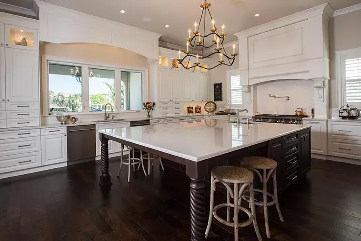 A kitchen with white cabinets , a large island , stools and a chandelier.