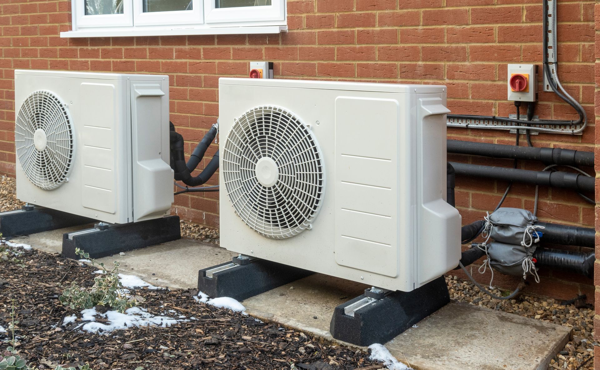 Two white outdoor air conditioning units beside a brick house with pipes and snow on the ground