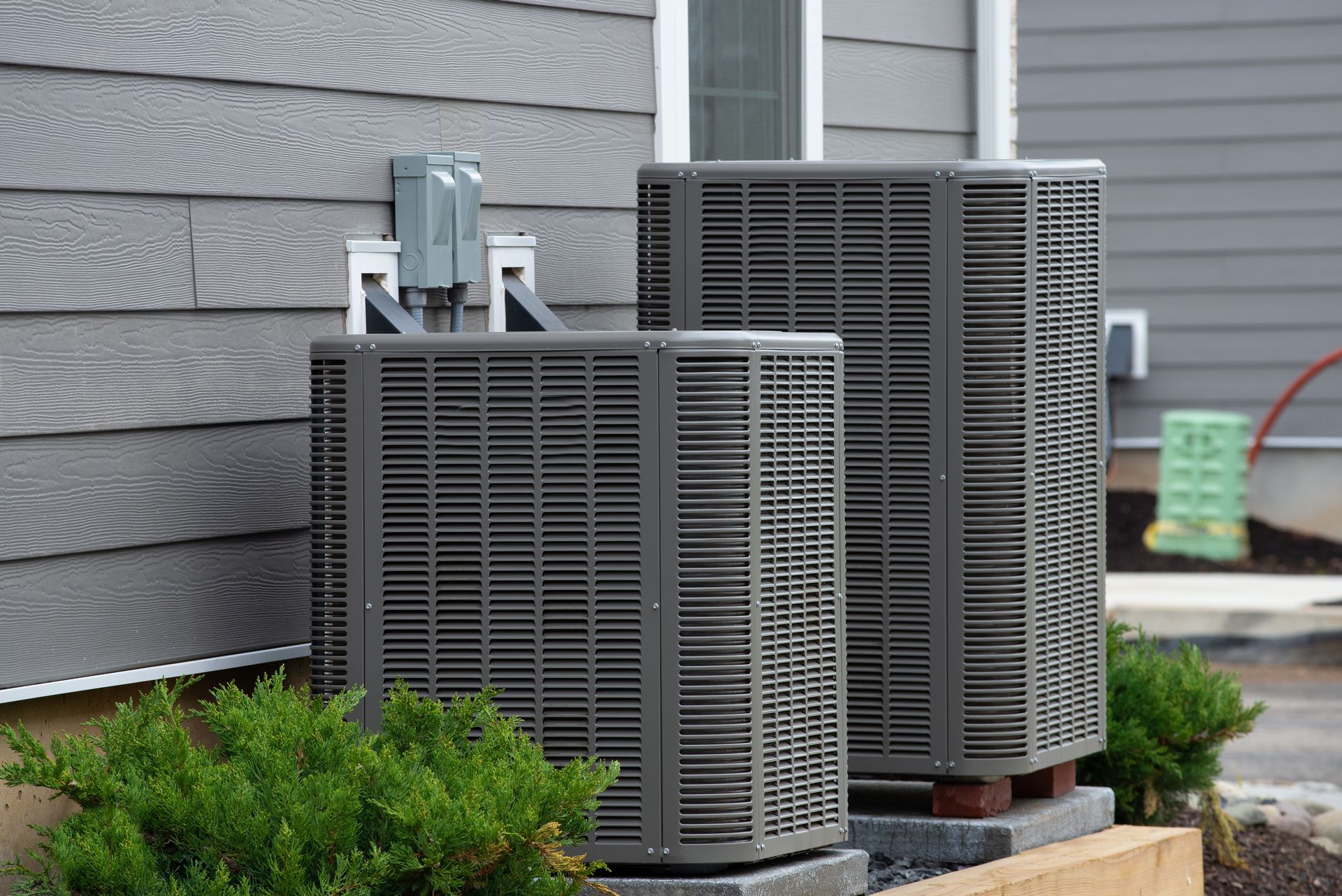 Two dark-gray HVAC condenser units sit on concrete pads outside a house with gray siding.