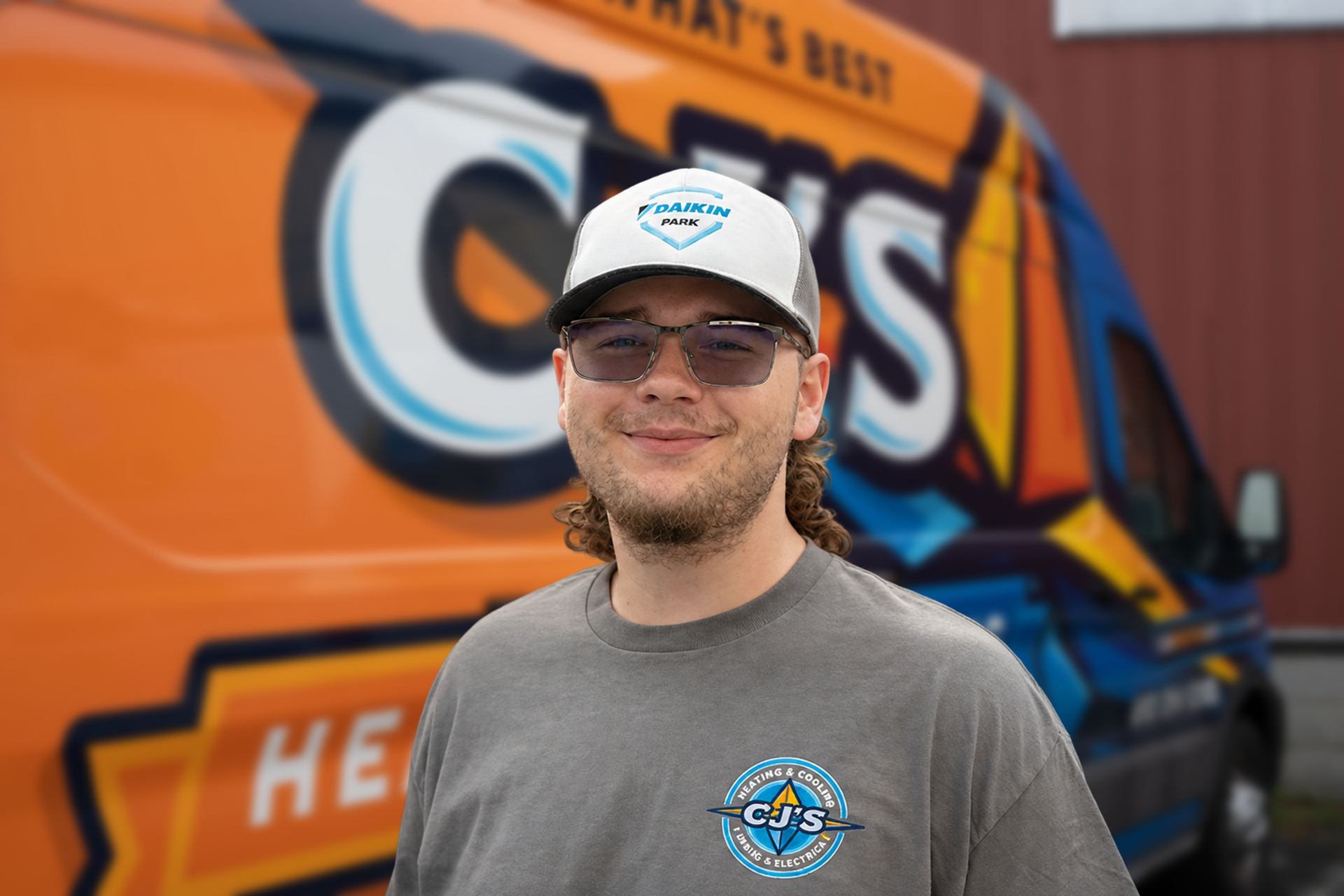 Man standing before an orange-and-blue food truck, wearing a white cap and gray shirt, smiling at the camera