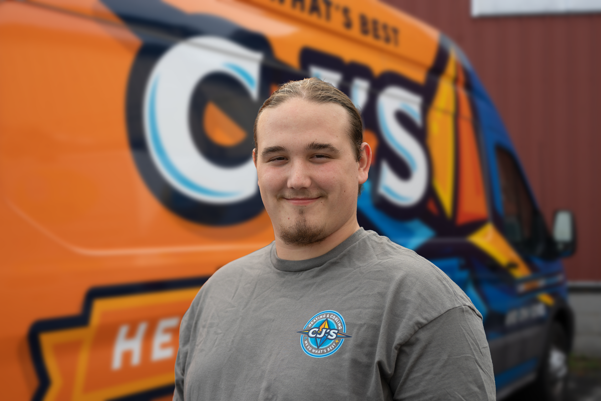 Smiling person in gray shirt standing before an orange service van with a large logo