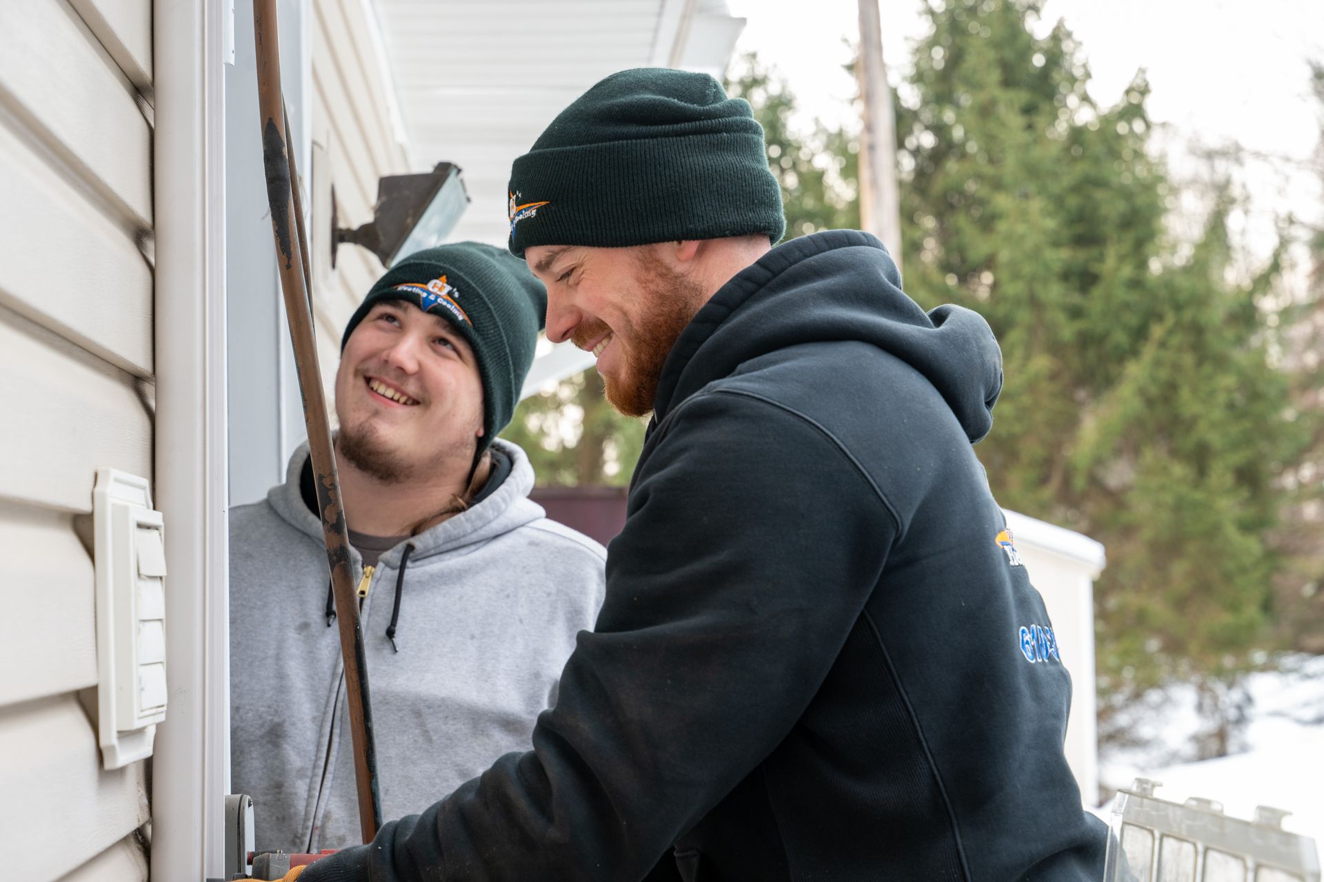 Two people working on a house exterior in winter, one wearing a beanie and smiling while holding a tool.