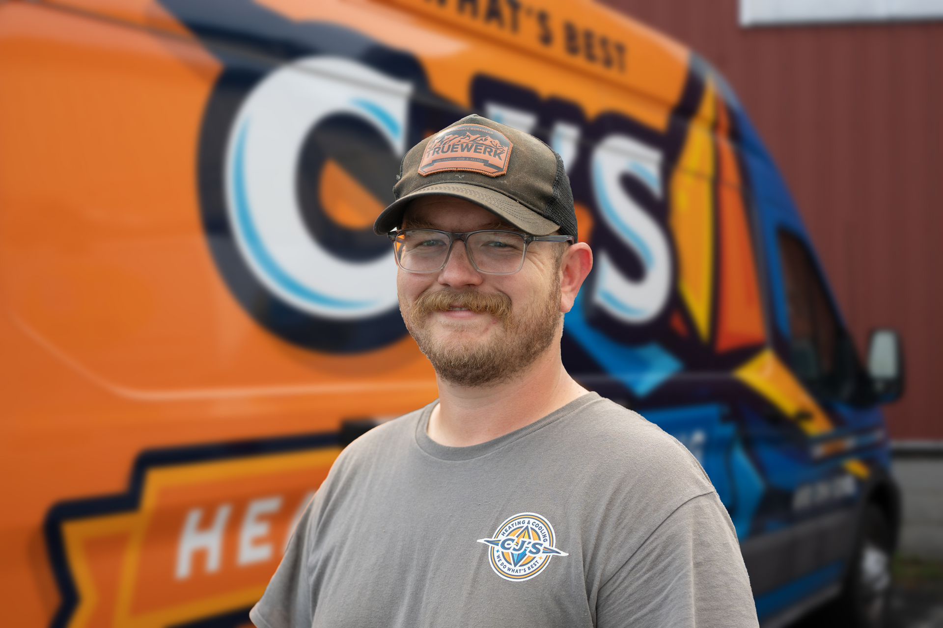 Smiling man in cap and glasses standing before an orange-and-blue service truck