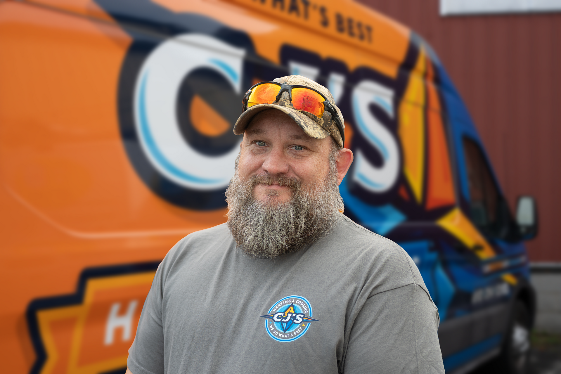 Man standing before an orange and blue truck, wearing a gray shirt, cap, and sunglasses on the brim
