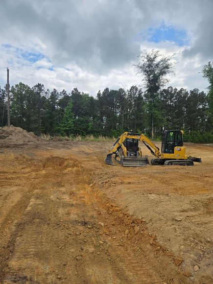 Yellow excavator on a dirt lot with trees and cloudy sky in the background.