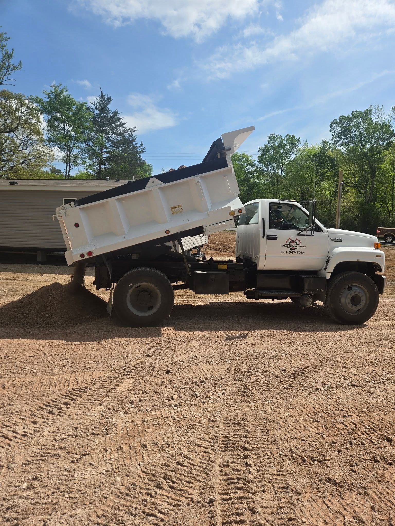 White dump truck raised at an angle on a gravel lot, with trees and blue sky in the background