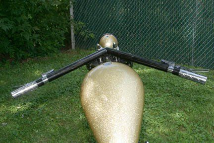 A motorcycle with a gold tank and black handlebars is parked in the grass.
