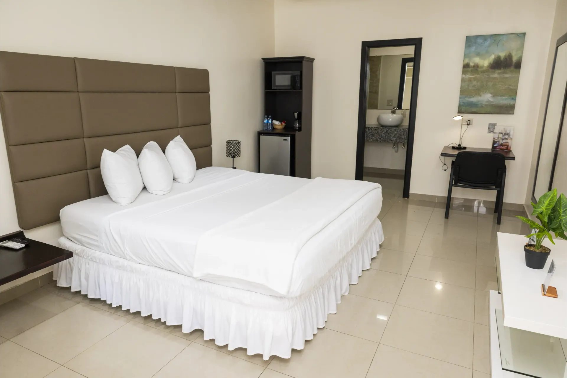 Bedroom with a white bed, beige headboard, mini-fridge, and a desk. A bathroom is in the background.