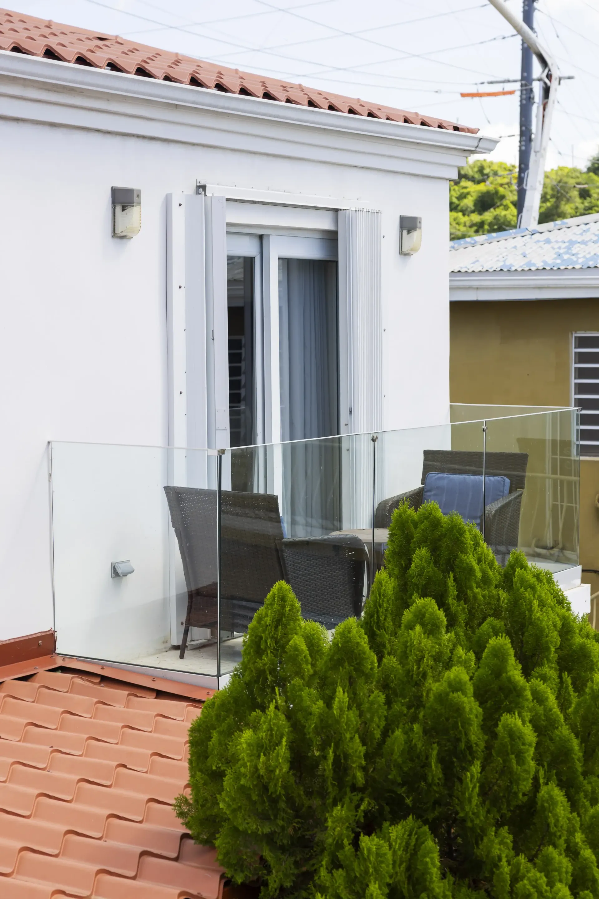 Balcony with glass railing, wicker furniture, and a green bush. Red tile roof and white building.