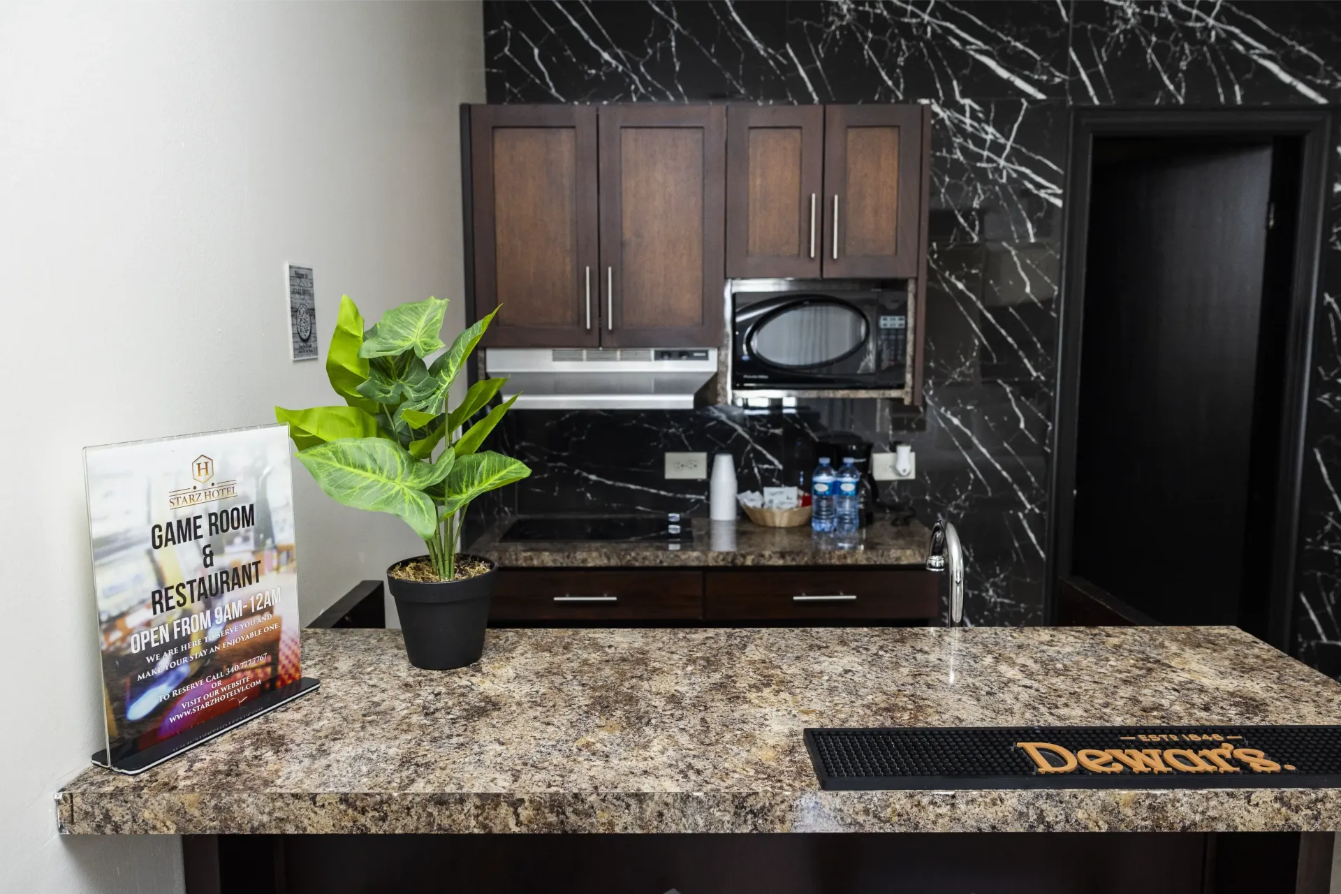A small kitchen area with a countertop, cabinets, and appliances. A plant and artwork sit on the counter.