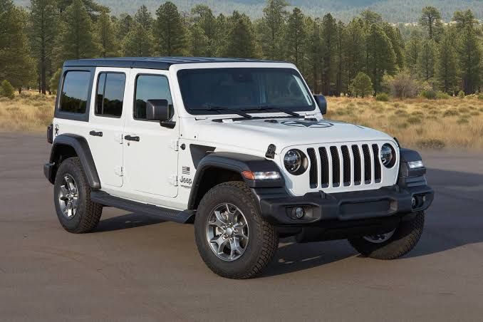 White Jeep Wrangler parked on dirt road, against a backdrop of trees and a bright sky.