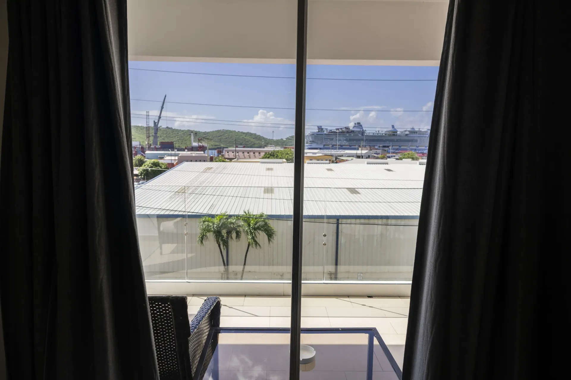 View from a room with curtains open onto a port with buildings, boats, and cranes under a blue sky.
