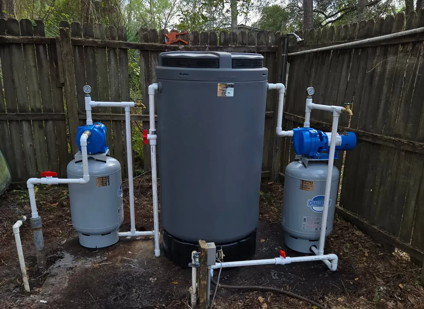 Outdoor water treatment setup with gray tank, two blue cylinders, and white pipes in a fenced yard
