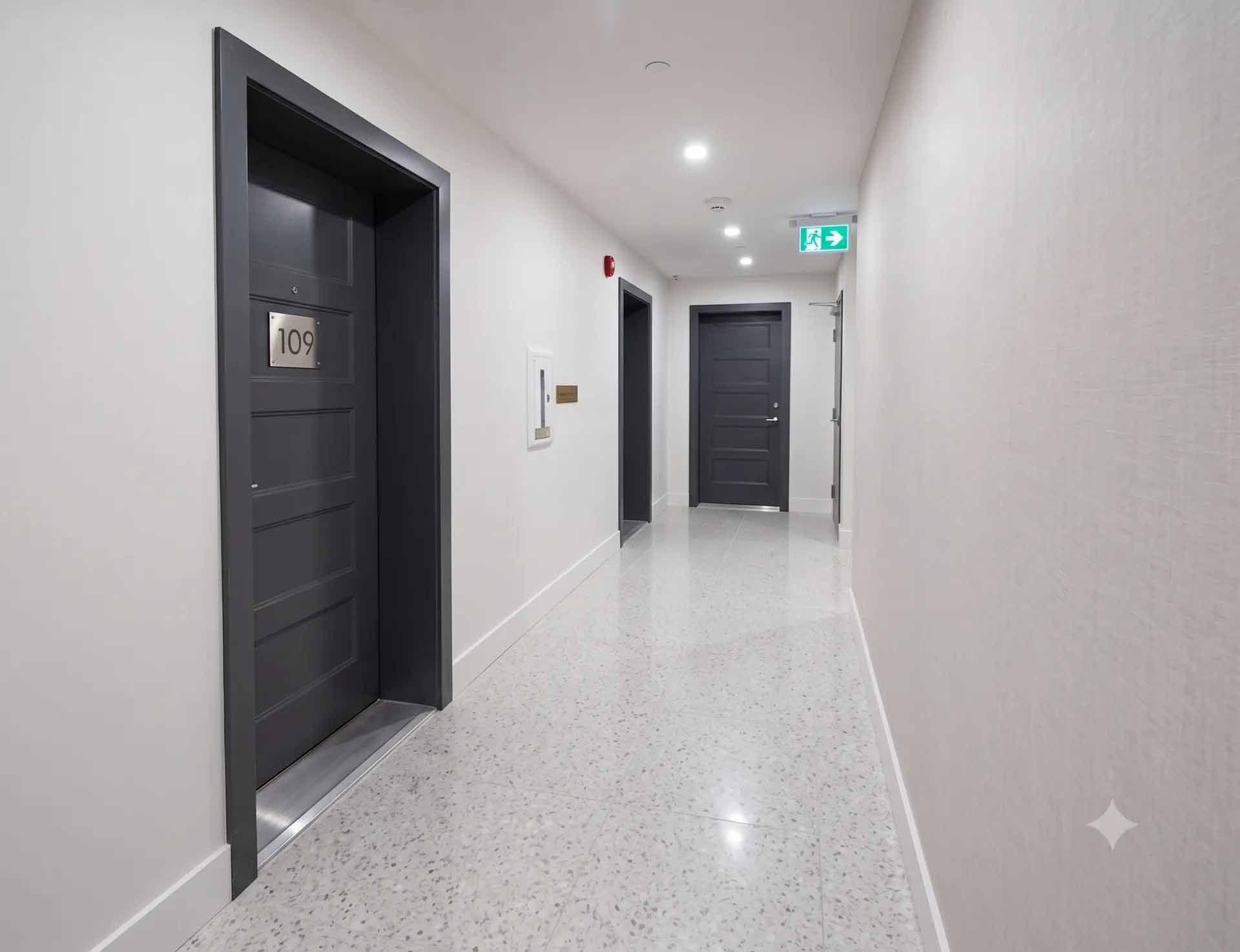 Couloir moderne avec portes grises, murs blancs et sol en terrazzo, éclairé par un panneau de sortie de secours
