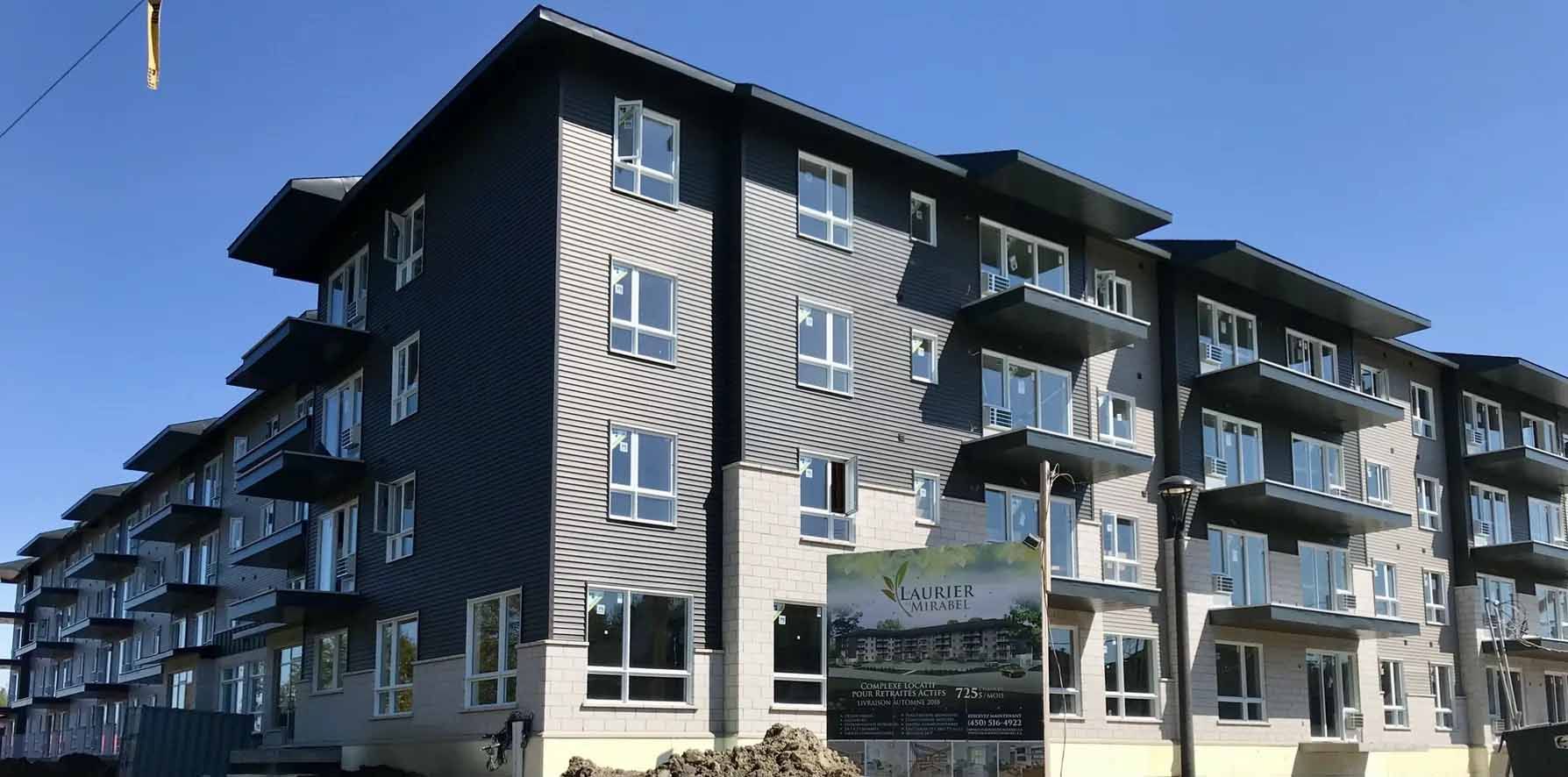 Complexe d'appartements moderne avec revêtement gris, balcons et entrée paysagée sous un ciel bleu