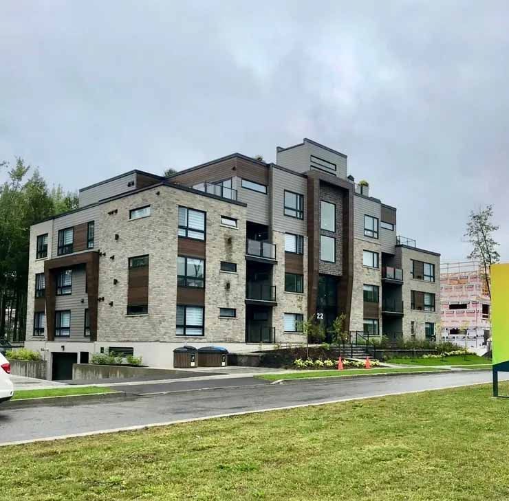 Immeuble d'appartements moderne avec façade en pierre et en bois sombre, jouxtant une pelouse sous un ciel nuageux.